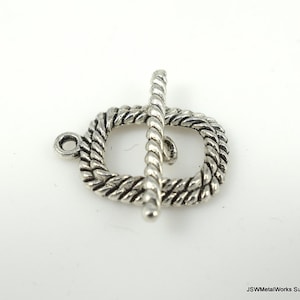 Op de afbeelding: Zilverkleurige metalen toggle sluiting met een touwontwerp. De sluiting is ovaalvormig met een rechte balk die door het midden gaat.