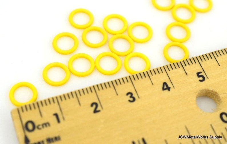 May include: A close-up of a ruler measuring yellow jump rings. The ruler is marked in centimeters and inches. The jump rings are scattered on a white surface.