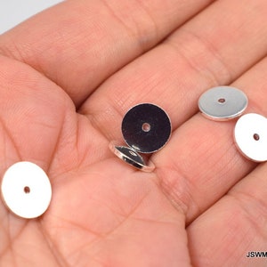 May include: Four silver metal round beads with a hole in the center. The beads are on a hand.