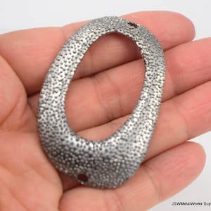 May include: A silver oval-shaped metal pendant with a textured surface and two holes at the top.