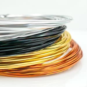 Könnte beinhalten: Nahaufnahme von mehreren Spulen Basteldraht in verschiedenen Farben. Die Farben umfassen Silber, Schwarz, Gold und Kupfer. Der Draht ist eng gewickelt und gestapelt, wodurch eine optisch ansprechende Darstellung entsteht. Der Hintergrund ist weiß.