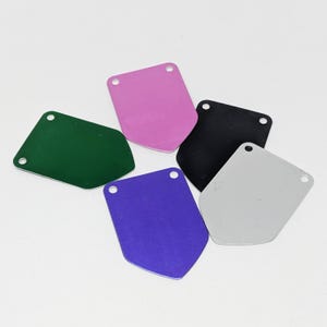 May include: Five blank metal tags in different colours: green, pink, black, purple, and silver. Each tag has two holes at the top for attaching a string or ribbon.