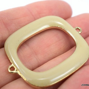 May include: A gold-toned metal frame with a cream-colored enamel finish. The frame has two small loops on the top and bottom for attaching to other jewelry or crafts.