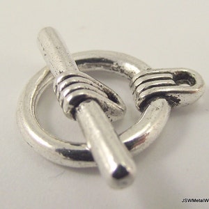 May include: Silver toggle clasp with a round ring and a straight bar. The ring and bar are connected by a decorative knot.
