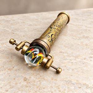 May include: A brass kaleidoscope featuring an intricate etched scrollwork pattern on the cylindrical body. A clear glass marble with swirling red, yellow, and black internal patterns is held in place by a metal bracket at the viewing end.