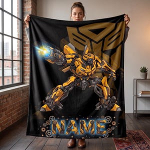 May include: A black fleece blanket featuring a yellow and grey robot character in a dynamic pose with a glowing blue cannon. The background displays a large gold Autobot logo, and the word NAME is printed at the bottom in a metallic font.