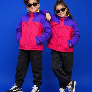 May include: Two matching puffer jackets featuring a vibrant colour-blocked design with bright pink main panels, deep purple shoulders and sleeves, and a high collar. Each jacket includes a front zip closure with snap buttons and black cargo trousers.
