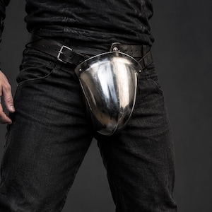 Puede incluir: Una coquilla de acero pulido sujeta a un cinturón de cuero negro, sobre unos vaqueros oscuros. El conjunto incluye botas de cuero marrón oscuro con vuelta y un protector de antebrazo de cuero a juego, creando un aspecto de protección.