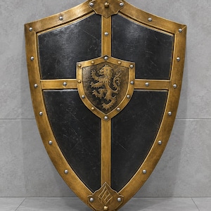 May include: A heater-style shield featuring a dark charcoal-colored surface with a metallic gold-toned frame and cross-shaped reinforcement. A central gold-toned crest displays a raised lion emblem, and the entire structure is accented with silver rivets.