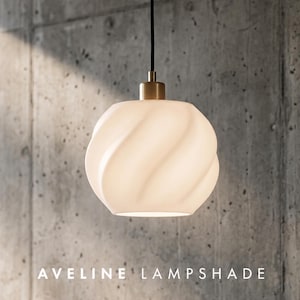 May include: A spherical white glass lampshade with a swirling, twisted ribbed texture. The shade is suspended by a black cord and features a metallic brass-toned fixture at the top. The text AVELINE LAMPSHADE is printed at the bottom.