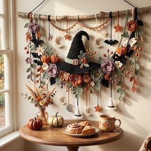 May include: A wall hanging featuring a central black crocheted witch hat surrounded by crocheted flowers, pumpkins, bats, stars, and moons suspended from a wooden branch. Below, a wooden table holds a vase of dried flowers, pumpkins, and a plate of pastries.