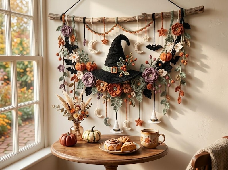 May include: A wall hanging featuring a central black crocheted witch hat surrounded by crocheted flowers, pumpkins, bats, stars, and moons suspended from a wooden branch. Below, a wooden table holds a vase of dried flowers, pumpkins, and a plate of pastries.
