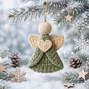 May include: A crocheted angel ornament featuring a sage green dress, a beige head, and beige wings. A small beige crocheted heart is centered on the chest. The ornament hangs from a simple tan string loop, showcasing detailed yarn texture.