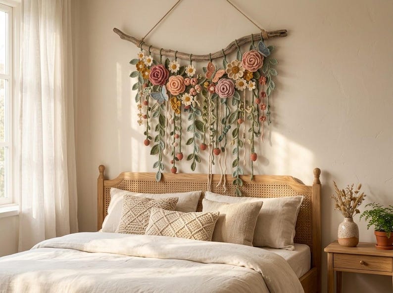 May include: A wall hanging featuring a curved piece of driftwood adorned with crocheted flowers in shades of pink, yellow, and white, along with green crocheted leaves, vines, and small butterflies, suspended by a simple rope cord.