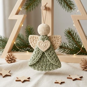 May include: A crocheted angel ornament featuring a round cream-colored head, sage green dress, and cream-colored wings. A small cream heart is centered on the chest. The ornament hangs from a thin string and is crafted with textured yarn.