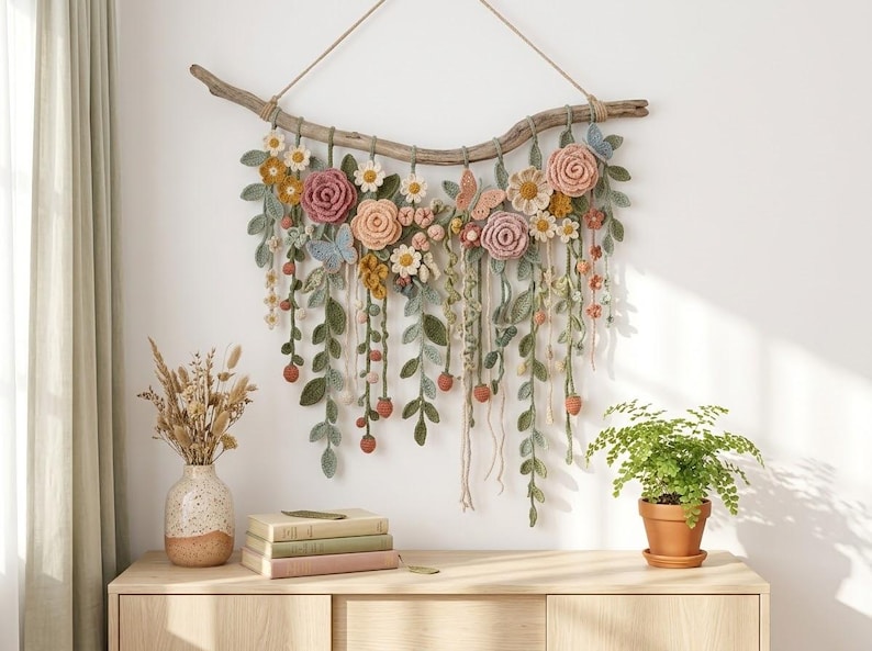 May include: A wall hanging featuring a piece of driftwood with crocheted flowers, leaves, and butterflies in shades of pink, yellow, green, and blue. The piece includes long, dangling vines with small crocheted berries hanging from the driftwood.