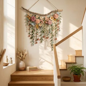 May include: A wall hanging featuring a horizontal wooden branch adorned with crocheted flowers in shades of pink, yellow, and white, interspersed with green crocheted leaves, vines, and small blue butterflies, suspended by a simple twine cord.
