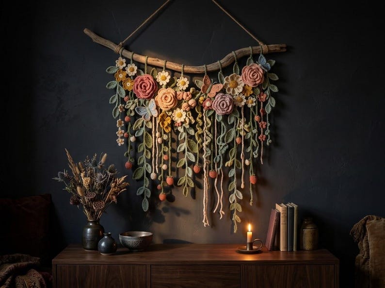 May include: A wall hanging featuring crocheted flowers, leaves, and butterflies in shades of pink, orange, yellow, and green, suspended from a natural wooden branch. The floral arrangement includes roses, daisies, and trailing vines with small berries.
