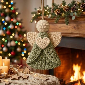 May include: A crocheted angel ornament featuring a round cream-colored head, cream-colored wings, and a sage green dress with a cream-colored heart centered on the chest. The ornament hangs from a thin jute twine loop for display purposes.