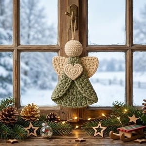 May include: A crocheted angel ornament featuring a sage green dress, cream-colored wings, and a matching cream-colored heart centered on the chest. The figure has a round cream-colored head and is suspended by a simple twine loop for hanging.