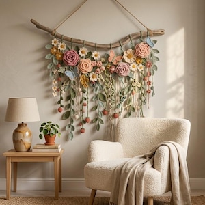 May include: A wall hanging featuring crocheted flowers, leaves, and butterflies in shades of pink, yellow, and green, suspended from a natural wooden branch with a rope. The floral arrangement includes roses, daisies, and trailing vines.