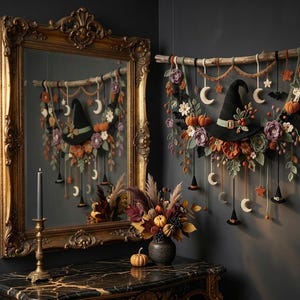 May include: A wall hanging featuring a black witch hat, crocheted flowers, pumpkins, and crescent moons suspended from a wooden branch. The display includes small hanging witch hats, stars, and bats, with a mirror reflecting the arrangement.