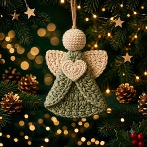 May include: A crocheted angel ornament featuring a round cream-colored head, cream-colored wings, and a sage green dress. A small cream-colored heart is centered on the chest of the angel. The ornament hangs from a thin, light brown twine loop.