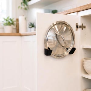 May include: A circular stainless steel pot lid held in place on the interior of a white cabinet door by two black plastic mounting brackets. The lid features a central metal handle and a polished reflective surface, securely stored for organisation.