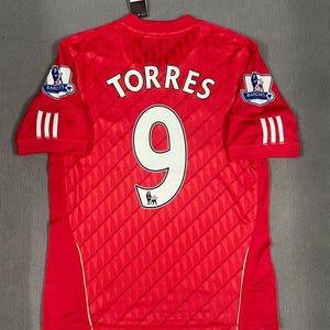 Puede incluir: Una camiseta roja de manga corta con un sutil patrón de diamantes, el nombre TORRES y el número 9 en blanco en la espalda. Las mangas muestran rayas blancas y parches de la Barclays Premier League. Una etiqueta negra está unida al cuello.