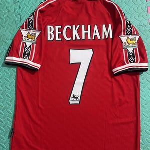 May include: A red short-sleeved jersey with a white collar and black trim. The back features the name BECKHAM in white letters above the number 7. Each sleeve displays a Champions 1998-1999 patch and an Umbro logo. An Umbro tag is attached.