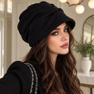 May include: A solid black knit beanie hat featuring a soft, slouchy silhouette with a structured brim. The hat is crafted from a textured, ribbed fabric that provides a gathered, layered appearance around the crown, offering a classic look.