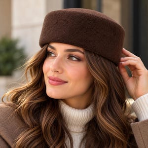 May include: A dark brown pillbox hat with a textured, felted surface. The hat features a flat top and a structured, cylindrical shape that sits firmly on the head, providing a clean and simple silhouette without any additional embellishments.