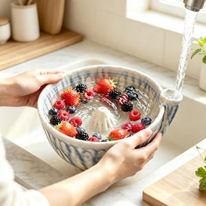 Puede incluir: Un cuenco de cerámica redondo con un patrón azul y blanco, que presenta un cono de drenaje central y un pico lateral. El cuenco está lleno de agua, fresas, frambuesas y moras frescas, mientras el agua fluye desde un grifo hacia el pico.