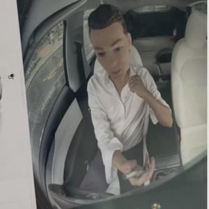 May include: A person with short brown hair wearing a white long-sleeved button-down shirt sits in the driver seat of a vehicle with light-colored interior upholstery. The person is holding a small metallic object in their hand while looking forward.