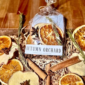 May include: A clear plastic bag containing dried orange slices, apple slices, cinnamon sticks, star anise, and rosemary sprigs. A white rectangular label on the front reads AUTUMN ORCHARD SIMMER POT POTPOURRI in black text. The contents rest on shredded paper.