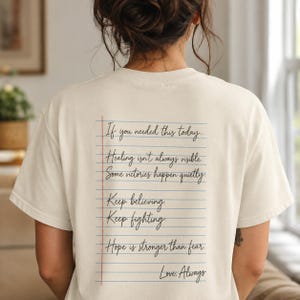 May include: A cream-colored t-shirt featuring a graphic on the back that resembles a sheet of lined notebook paper. The text reads: If you needed this today... Healing isn't always visible. Some victories happen quietly. Keep believing. Keep fighting. Hope is stronger than fear. Love, Always.