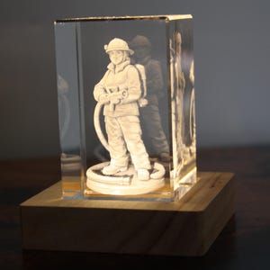 Puede incluir: Un bloque de vidrio rectangular transparente con una imagen 3D grabada con láser de un bombero en uniforme sosteniendo una manguera. El bloque de vidrio está montado sobre una base de madera cuadrada que proporciona iluminación.