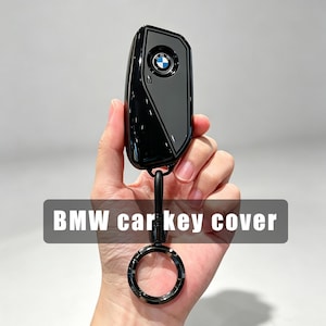 May include: A glossy black BMW car key cover featuring a central circular BMW logo emblem. The cover is attached to a black metal ring keychain. The text "BMW car key cover" is displayed in white letters on a dark gray rectangular background.