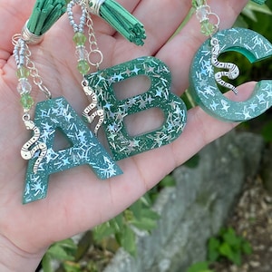 May include: Three green resin letter charms with silver glitter and a silver snake charm. Each charm has a silver chain and a green tassel.