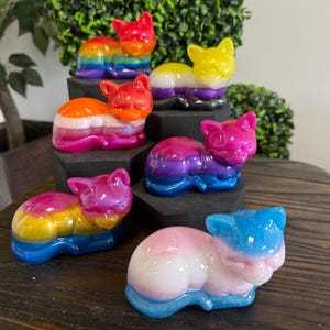 May include: A collection of six small, smooth, resin cat figurines in various color combinations. Each cat is depicted in a resting, curled-up position with ears pointed upward. The colors include rainbow, yellow-white-purple, orange-white-pink, pink-purple-blue, yellow-pink-blue, and pink-white-blue gradients.