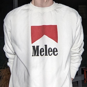 May include: A white sweatshirt with a red and white graphic design and the word "Melee" in black. The sweatshirt is worn by a person standing outside at night.