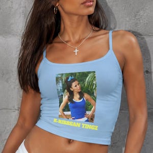 May include: A light blue ribbed crop top featuring a rectangular graphic print of a person wearing a blue tank top with the yellow text K-RIBBEAN TINGZ printed underneath the image. The garment has thin shoulder straps and a scoop neckline.