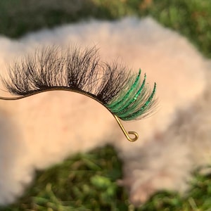 May include: A pair of false eyelashes featuring a curved black band with long, dark, wispy fibers on the left side and vibrant green, glitter-infused fibers on the right side, held by a thin, gold-toned metal wire applicator tool.