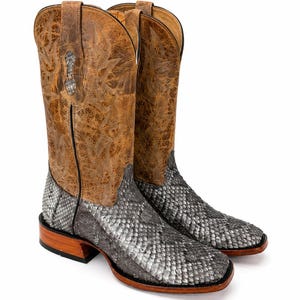May include: A pair of western-style boots featuring a textured grey snakeskin-patterned foot and a tall, tan-colored shaft with intricate stitching details. The boots have a square toe design, a stacked wooden heel, and pull tabs at the top.