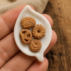 May include: A small, white, leaf-shaped ceramic dish holding four miniature brown cookies. The cookies have distinct textures, including a ridged oval, a circular ring shape, a textured round piece, and a small pretzel-like shape with holes.