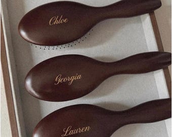 May include: Three dark brown oval hairbrushes with smooth wooden handles. Each brush features a unique name engraved in a gold script font on the back: Chloe, Georgia, and Lauren. The brushes are arranged vertically in a row.