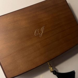Puede incluir: Una caja rectangular de madera con un acabado liso en color marrón medio. La tapa presenta las iniciales O.J. grabadas en color dorado con letra cursiva. Un cierre de metal dorado está sujeto en la parte frontal, adornado con una borla de tela negra.
