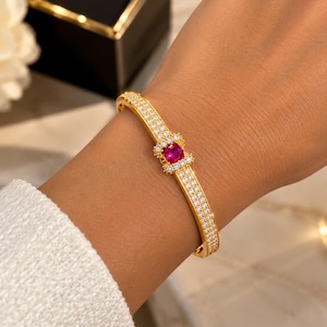 Peut inclure: Un bracelet jonc articulé doré orné de deux rangées parallèles de petites pierres pavées transparentes. Le centre du bracelet présente une pierre précieuse rose vif de taille carrée entourée d'un halo de petites pierres pavées.