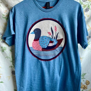 May include: A heather blue short-sleeve t-shirt featuring a large circular patch on the chest. The patch displays a stylized duck composed of various floral and patterned fabric segments in shades of blue, pink, and dark red, with cattails.