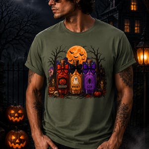 May include: An olive green t-shirt featuring a graphic print of three glass bottles labeled Bourbon, Whisky, and Gin. The bottles are decorated with bows and Halloween-themed icons including a pumpkin, a ghost, and a spider, set against a full moon.